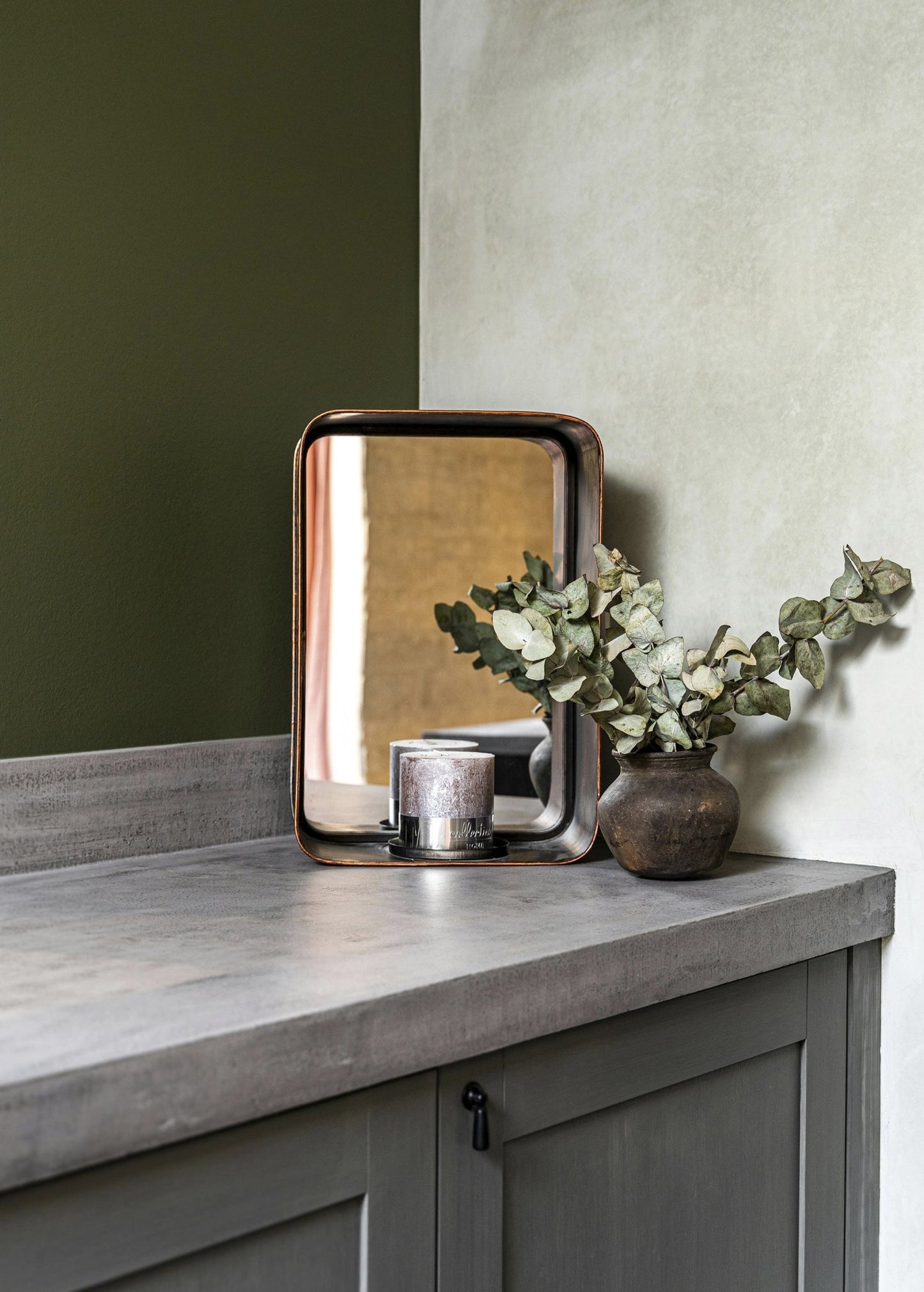 Decorative setup with eucalyptus stems in a rustic vase, candle, and mirror on a grey countertop, ideal for showcasing home fragrance accessories.